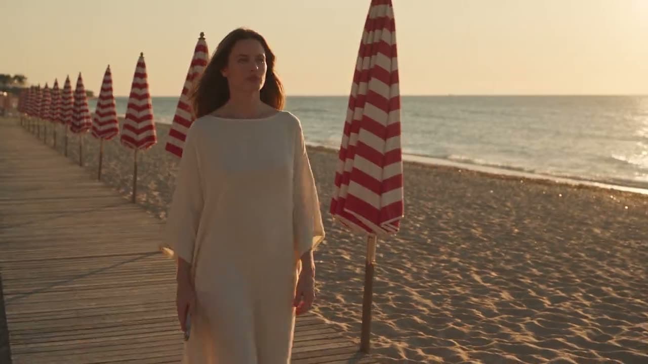 A woman walks a wooden beach boardwalk at golden hour past rows of striped parasols, the Mediterranean behind her, her phone lit in her hand.